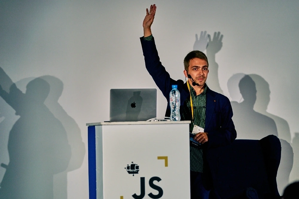 Person presenting on stage at a JS conference with one hand raised.