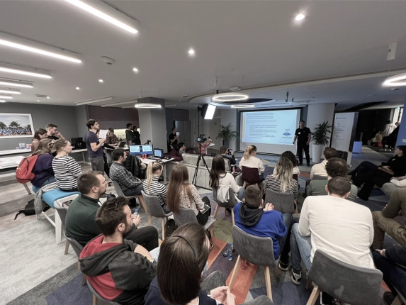 Attendees seated in a modern conference room, watching a presenter in front of a screen.