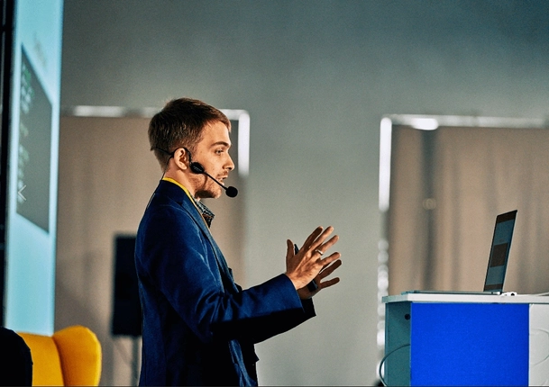 Presenter speaking on stage with a microphone headset and using a laptop.