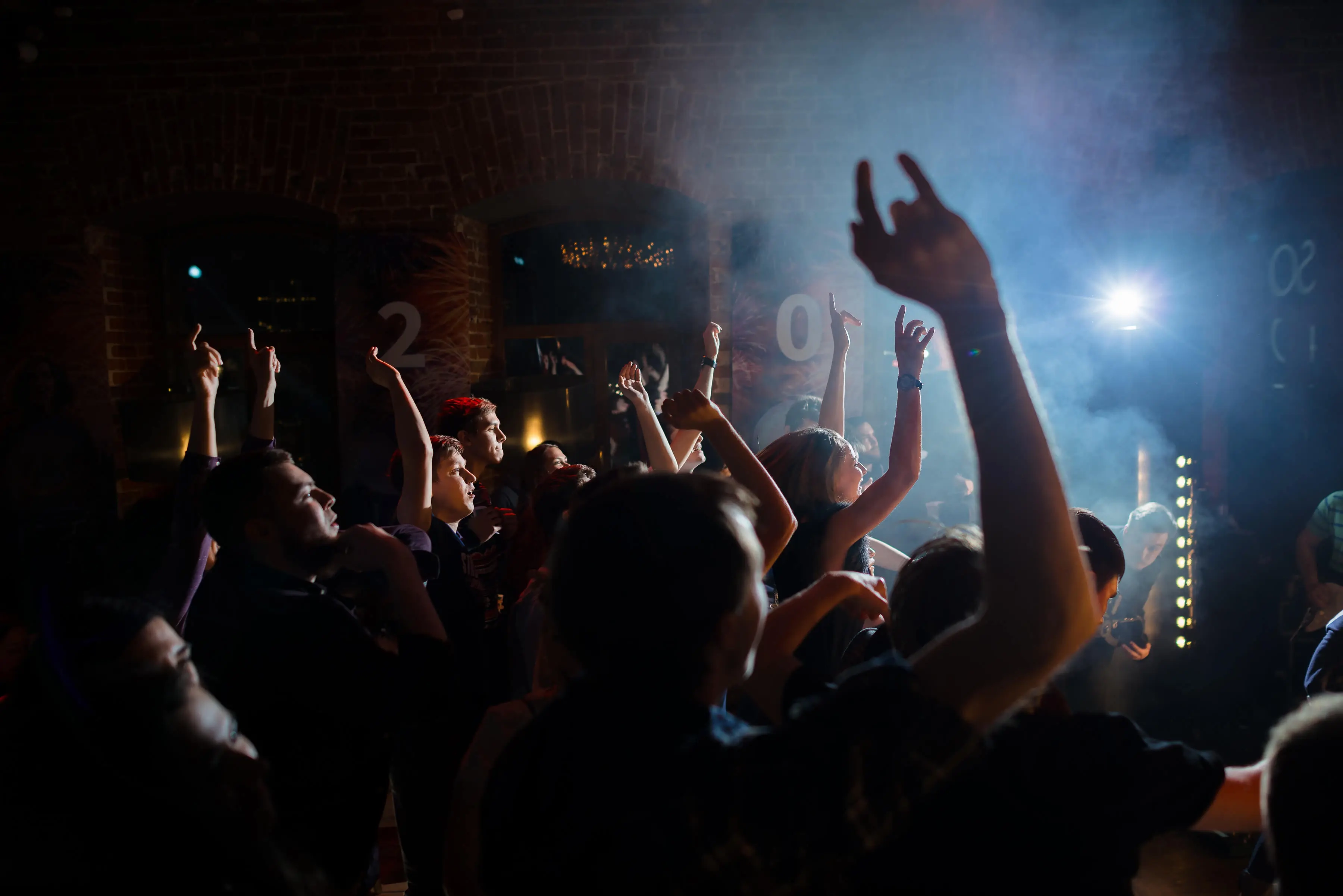 People dancing and raising their hands at a lively event with vibrant lighting and a smoky atmosphere.