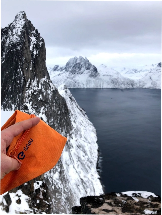Hand holding an orange GridU item with a snowy mountain landscape and fjord in the background.